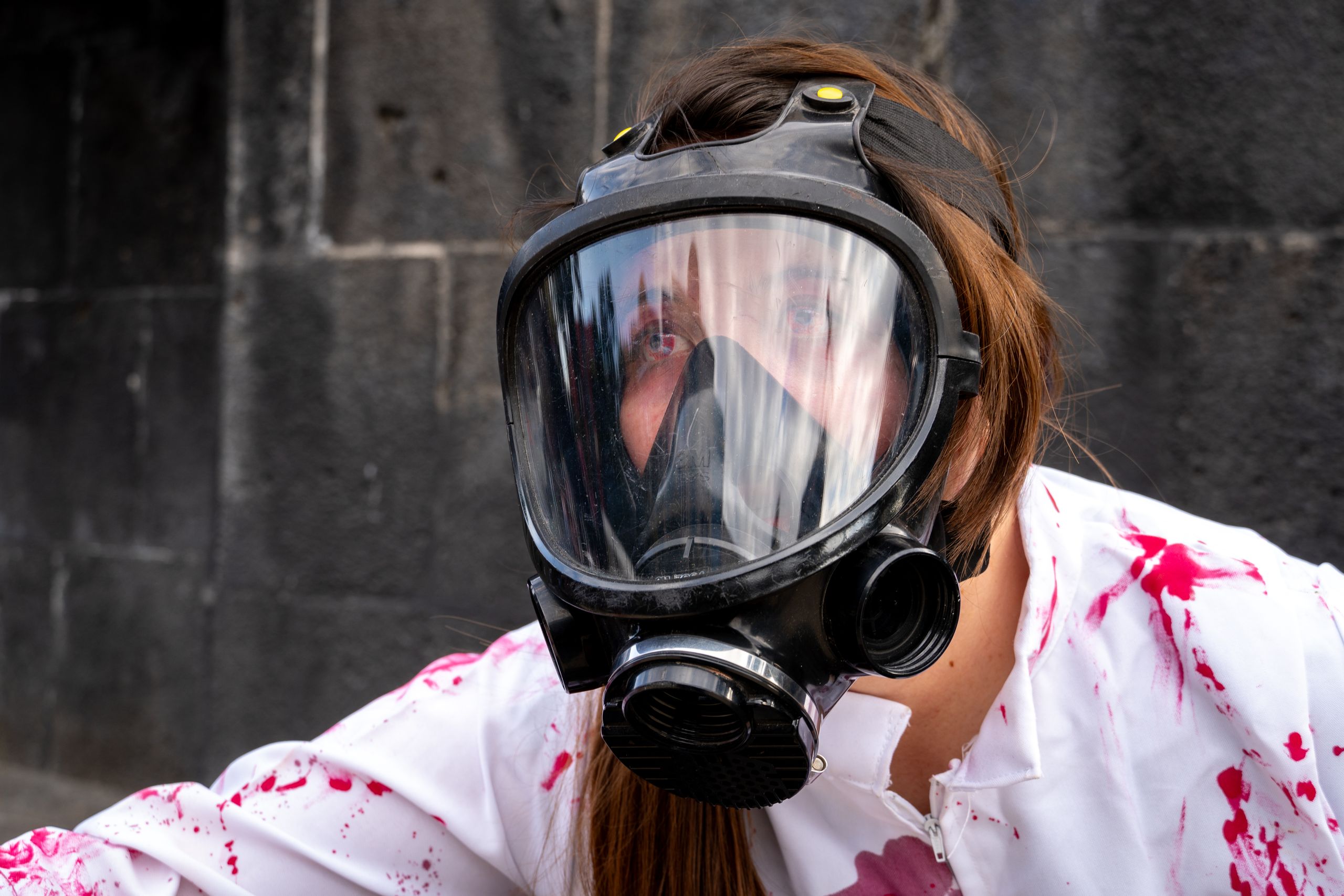 Close-up of a person wearing a gas mask and bloodstained white outfit outdoors.